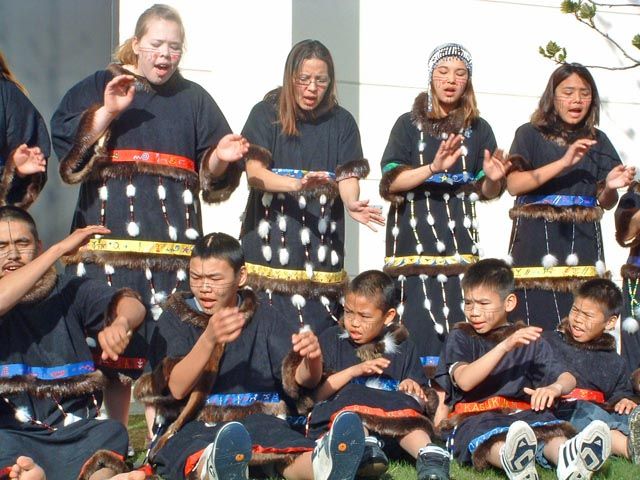 A group of young people , girls standing, boys seated, dance in Alutiiq regalia.