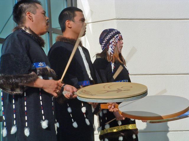 Three people beating large, circular, hand held drums.