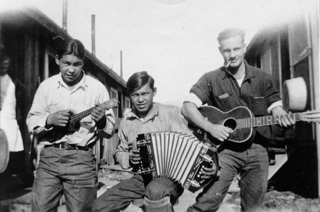 Three men outdoors holding instruments - a ukulele, accordion, and guitar.